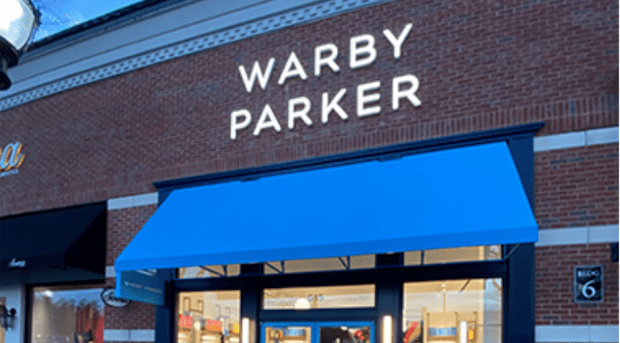 Storefront of a Warby Parker retail location with a blue awning and illuminated sign on a brick facade.